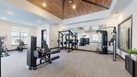 Located in Rosemount, MN, this fitness center offers a well-appointed, single-floor exercise space designed with both functionality and modern aesthetics in mind. The room's most striking architectural feature is its soaring vaulted ceiling clad in warm wood paneling, complemented by recessed lighting that creates a bright and energizing atmosphere. Large, black-framed windows line the walls, flooding the interior with natural light and providing views of the surrounding area. The flooring throughout is a neutral, textured carpet that adds comfort underfoot while maintaining a clean, contemporary look. Full-length mirrors are strategically placed to visually expand the space and support proper form during workouts. The equipment selection is comprehensive, including True and Spirit branded cardio machines such as treadmills, stationary bikes, and rowing machines, alongside a robust lineup of strength training equipment including cable machines, a power rack, and various selectorized strength machines. A dedicated free weights area features a well-organized rack stocked with kettlebells, medicine balls, dumbbells, resistance bands, and a stability ball. The entrance is framed by sleek glass-paneled double doors, lending the space a polished, professional feel. Overall, this fitness center delivers a well-rounded workout environment in a clean, modern setting.
