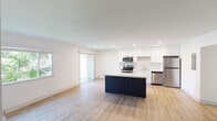 This single-floor unit offers a clean, modern aesthetic defined by an open-concept layout that seamlessly connects the kitchen and living areas. Light hardwood flooring runs throughout the main living spaces, enhancing the bright and airy atmosphere created by recessed lighting and multiple windows that frame lush greenery outside.

The kitchen is a standout feature, equipped with a full suite of stainless steel appliances including a refrigerator, range, microwave, and dishwasher. Crisp white cabinetry extending to the ceiling provides ample storage, while a tile backsplash adds a subtle textural detail. A centrally positioned island with a dark navy base and light countertop serves as both a functional workspace and a visual anchor for the open living area. A sliding glass door offers access to the outdoors, further connecting the interior to the surrounding environment.

The bedrooms are carpeted for comfort and receive natural light through well-placed windows. Bedroom closets feature mirrored sliding doors, adding depth and practicality. The bathrooms are finished in a clean, neutral palette — one includes a shower stall, while another offers a full shower/tub combination. The overall design prioritizes functionality and modern simplicity throughout.