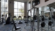 This fitness center is designed to support a comprehensive workout experience within a single, open-plan space. Floor-to-ceiling windows along one wall flood the room with abundant natural light while offering views of the surrounding greenery and outdoor landscape, creating an energizing atmosphere for exercise. The soaring ceiling height enhances the sense of openness and airiness throughout the space. The flooring features a durable, wood-look surface well-suited for high-traffic fitness use. The facility is outfitted with multiple treadmills positioned along the windowed wall, allowing users to enjoy the outdoor views while engaging in cardio exercise. A cable weight machine and a dedicated free-weight rack with a workout bench provide ample strength training options. A wall-mounted flat-screen television adds an entertainment element to the space. Full-length mirrors are strategically placed to expand the visual depth of the room and assist users in monitoring their form. The overall design is clean and contemporary, with a neutral color palette and modern finishes that contribute to a focused and motivating fitness environment.
