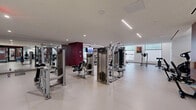Located at 42 Broad St W in Mount Vernon, NY, this single-floor fitness center offers a generously sized exercise space designed to support a full range of workout activities. The facility is thoughtfully organized into distinct functional zones, providing a comprehensive fitness experience.

The cardio area features a row of treadmills and stationary bikes, including Spinning cycles, positioned alongside large windows that allow natural light to fill the space and offer views to the outdoors. Bold burgundy accent walls add a striking visual element throughout, contrasting with the clean white walls and light-toned resilient flooring.

A well-equipped strength training zone houses multiple cable and selectorized weight machines, free-weight benches, and resistance equipment arranged to maximize floor space and ease of movement. An additional open area provides room for functional training, with a dumbbell rack and wall-mounted display screen, ideal for group classes or solo routines.

A separate studio space features floor-to-ceiling mirrors and ballet-style wall-mounted barre rails, suitable for dance, yoga, or stretching sessions. Glass sliding doors connect this studio to the main gym floor, maintaining an open, integrated feel. The facility is lit by a combination of recessed ceiling lights and linear pendant fixtures, creating a bright and energizing atmosphere throughout.