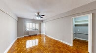 This efficiently designed single-floor unit offers a thoughtful layout comprising a living room, kitchen, bathroom, entrance, and two closets. The living room is the centerpiece of the space, featuring striking warm-toned parquet wood flooring laid in a classic basket-weave pattern that reflects light beautifully throughout the room. Neutral gray walls provide a clean, versatile backdrop, while a ceiling fan with integrated lighting adds both comfort and function. A wide window allows natural light to fill the room and provides views of the surrounding building exterior. A baseboard heating unit runs beneath the window, ensuring year-round comfort. A graceful arched doorway connects the living area to the rest of the unit, adding an architectural detail that softens the overall aesthetic. The kitchen is compact yet practical, outfitted with white shaker-style cabinetry, dark countertops, a gas range, a full-size refrigerator, and a single-basin sink, all set atop light-toned tile flooring. The bathroom maintains a clean, classic look with white mosaic tile flooring, white wainscoting, a pedestal-style vanity with a mirror, and a shower-tub combination. Two closets provide ample storage throughout the unit.