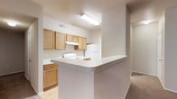 This single-floor residence offers a functional and well-organized layout designed for comfortable everyday living. The kitchen serves as a central feature of the space, showcasing warm honey-toned wood cabinetry that lines the walls and provides ample storage. A peninsula counter extends outward, creating a natural partition between the kitchen and the adjoining living area while offering additional prep and casual dining space. White appliances, including a freestanding electric range and a full-size refrigerator, are neatly integrated into the layout. A range hood is positioned above the stove, and recessed ventilation ensures proper airflow throughout the cooking area. Overhead fluorescent lighting illuminates the kitchen with bright, even light. Light-toned countertops complement the cabinetry and contribute to the clean, neutral aesthetic carried throughout the space. The flooring transitions from hard surface in the kitchen to plush carpeting in the adjacent living area, adding warmth and comfort underfoot. The overall color palette of soft beiges and warm neutrals creates a cohesive and inviting atmosphere. A hallway visible from the main living area suggests additional rooms beyond, rounding out the practical and straightforward design of this single-level home.