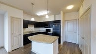 This single-floor space showcases a well-appointed kitchen designed for both functionality and style. The kitchen is anchored by a central island with a light granite countertop and white base, providing additional workspace and casual dining potential. Pendant lights hang above the island, adding focused illumination and a contemporary touch.

The cabinetry throughout is finished in a rich, dark tone, creating a bold contrast against the light granite countertops that extend along the perimeter walls. A full suite of black appliances, including a refrigerator, range, microwave, and dishwasher, complements the dark cabinetry seamlessly. A built-in wine rack adds a refined detail above the refrigerator.

The sink is positioned along the side wall, offering practical workflow within the L-shaped layout. Hardwood-style flooring in a warm medium tone runs throughout, adding warmth and continuity to the space. Neutral walls and flush ceiling lighting keep the overall aesthetic clean and bright. A doorway visible from the kitchen suggests a smooth connection to adjacent areas of the home.