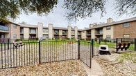Located along Preston Road in Dallas, Texas, this outdoor amenity space serves as a dedicated dog park within a multi-building residential community. The grounds are enclosed by a sturdy black iron fence with a gated entry, providing a secure environment for pets and their owners.

The park features a generously sized open lawn area with a mix of grassy patches and natural ground cover, offering ample room for pets to roam freely. Several pieces of pet agility equipment are distributed throughout the space, including tunnel obstacles and ring features, encouraging active play. Seating is well-considered, with multiple benches — including a low wooden platform bench and a classic iron-and-wood park bench — positioned throughout for resident use.

Mature oak trees provide natural shade and a canopy overhead, contributing to a comfortable outdoor atmosphere across seasons. The surrounding two-story residential buildings feature a combination of brick and siding exteriors with covered balconies and chimneys, framing the park on multiple sides and creating a cohesive, community-centered feel.

Additional amenities visible within and adjacent to the space include waste disposal stations and a picnic table, making it a fully functional and thoughtfully equipped outdoor destination for residents and their pets at this Dallas community.