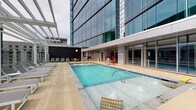 Located at 511 Fayetteville St in the heart of downtown Raleigh, NC, this rooftop amenity level offers an expansive outdoor retreat set against the backdrop of a striking glass tower. The space is defined by polished concrete throughout and seamlessly blends indoor and outdoor living via floor-to-ceiling glass walls and wide sliding doors.

A central outdoor pool anchors the space, flanked by rows of lounge chairs ideal for relaxing. Adjacent to the pool, a covered pergola area provides shade over casual seating and a bar or grill station, making it well-suited for entertaining. Nearby, a generous open-air dining zone features long communal tables with modern wire chairs, offering ample room for group gatherings.

A separate open lounge terrace showcases panoramic views of the Raleigh skyline, furnished with contemporary rounded seating clusters and low wood-plank benches. Raised planter boxes with lush greenery add a touch of nature to the urban setting, while sleek cylindrical columns frame the perimeter, reinforcing the building's modern architectural aesthetic.

The indoor-outdoor flow is enhanced by transparent glass partitions that allow city views to permeate throughout. This thoughtfully designed rooftop destination provides a dynamic mix of recreation, dining, and relaxation amenities in a sophisticated urban setting.
