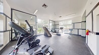 This well-appointed fitness center offers a generously sized, single-floor layout divided into distinct functional zones, providing a comprehensive workout environment. The space is designed with a clean, modern aesthetic, featuring white walls accented by bold dark trim that lends a structured, professional character throughout.

The cardio zone benefits from an abundance of natural light, with large multi-pane windows offering views of surrounding greenery. Equipment in this area includes treadmills, stationary bikes, elliptical machines, rowing machines, and a freestanding stair-climber unit. Full-length mirrors are strategically placed to enhance the sense of openness and allow users to monitor their form.

A dedicated strength training area transitions into a darker, more energetic atmosphere, characterized by a deep-toned ceiling and a striking, colorful mural with bold graffiti-style lettering. This zone is equipped with power racks, barbells, weight plates, cable machines, and functional training accessories. Rubber flooring throughout the entire facility provides durability and impact absorption.

The entry area features hardwood-style flooring and high ceilings with clerestory windows, creating a bright, welcoming arrival experience. Recessed and linear fluorescent lighting supplements natural light across all zones, ensuring consistent illumination for all-hours use.