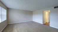 This single-floor space presents a straightforward and functional layout centered around a generously sized main room. The room is finished with wall-to-wall beige carpeting that adds a sense of warmth underfoot, while the neutral-toned walls provide a clean, understated backdrop suitable for a variety of interior styles. The textured popcorn ceiling adds a classic touch to the space, complementing the simple, unadorned walls lined with white baseboards that offer a crisp contrast to the soft flooring. Electrical outlets are conveniently positioned along the walls, supporting practical use of the space. A doorway visible along one wall leads to an adjacent area, suggesting additional rooms beyond the main living space, with a glimpse of hardwood-style flooring transitioning into the connected space. The open, empty layout of the main room allows for flexible furniture arrangement and use. Overall, the space carries a calm, neutral ambiance that serves as a solid canvas for personalization, making it well-suited for a range of occupants and purposes.