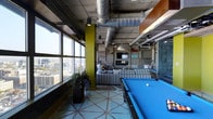 800 Tower City Apartments offers a striking communal lounge space defined by its bold industrial-chic aesthetic. The single-floor layout showcases exposed concrete ceilings with large-scale ventilation ductwork running overhead, lending an urban, loft-like character to the environment. A vivid yellow-green structural column anchors the room visually, complementing the eclectic mix of textures and materials throughout.

At the heart of the space sits a vibrant blue billiard table, making it a natural focal point for social gatherings. Adjacent to it, a generously sized seating area features striped upholstered sofas arranged around a fireplace, creating a warm and inviting atmosphere. A wall-mounted television and decorative signage — including a prominent "800" marker — add personality to the lounge backdrop, which is finished with rustic wood paneling for contrast.

Floor-to-ceiling windows line one entire wall, flooding the space with abundant natural light and offering sweeping panoramic views of the surrounding cityscape. Geometric patterned flooring adds a dynamic visual element underfoot, while potted greenery softens the industrial edges. Track and pendant lighting complete the layered ambiance, making this lounge equally suited for lively entertainment or relaxed socializing.