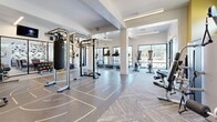 Located at 1850 SE 18th Ave in Ocala, Florida, this fitness center offers a comprehensive workout environment spread across a single, generously sized floor. The space is divided into two distinct zones that together provide a full range of exercise options for all fitness levels.

The cardio area is lined with modern treadmills and elliptical machines positioned along large, black-framed windows that fill the room with natural light and offer views of the surrounding outdoor greenery. Ceiling fans and recessed linear lighting ensure the space remains comfortable and well-lit throughout.

The functional training zone features a multi-station cable and pull-up rig, gymnastics rings, battle ropes, a heavy punching bag, and a full rack of colorful kettlebells and medicine balls. Floor markings on the durable training surface designate workout lanes and agility areas, accommodating high-intensity interval and circuit training. Large mirrors line the walls, enhancing the sense of openness. A dedicated free weights area with dumbbells and benches rounds out the strength training offerings.

The overall ambiance is bright and modern, with a neutral white and gray palette accented by bold yellow walls in the strength zone. Glass partitions provide visual connectivity to an adjacent lounge area, contributing to an inviting and motivating atmosphere throughout the facility.