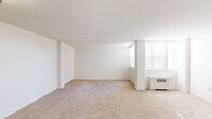 Located at 3701 Conshohocken Ave in Philadelphia, PA, this efficiently designed single-floor unit offers a clean, neutral aesthetic throughout. The open living area is finished with wall-to-wall beige carpeting and bright white walls, creating a light and airy atmosphere. A large window fitted with vertical blinds allows natural light to fill the space, while a wall-mounted HVAC unit provides climate control. The layout flows naturally from the living area toward the entryway, which features a dedicated closet for additional storage. The kitchen is well-appointed with white cabinetry extending to the ceiling, offering generous storage capacity. Dark laminate countertops provide a contrasting work surface, and the kitchen is equipped with a white gas range, an overhead ventilation hood, and a stainless steel sink. The full bathroom is finished with light tile flooring and white walls, and includes a toilet and a standard soaking tub with a shower rod. The overall design of the unit is clean and functional, with a consistent white palette that enhances the sense of space and keeps the interior feeling open and manageable throughout.