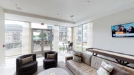This contemporary game room is designed to be a vibrant social hub, blending comfort and entertainment in a sleek, modern setting. The space is flooded with natural light through expansive floor-to-ceiling windows and glass doors that line the perimeter, offering unobstructed views of the outdoor pool and courtyard beyond. Sheer white curtains frame the windows, softening the abundant sunlight while maintaining the airy, open atmosphere.

The interior features a comfortable lounge area anchored by a large taupe sofa adorned with accent pillows and paired with rounded dark accent chairs, all arranged atop a plush area rug. A game table — ideal for billiards or air hockey — is positioned nearby, making this space a natural gathering spot for recreation and relaxation. High bar-height seating near the windows provides additional spots to enjoy the outdoor views.

The clean white ceilings and neutral-toned walls give the room a polished, contemporary feel, while the smooth flooring adds to the refined aesthetic. French-style glass doors lead directly to the outdoor amenity area, seamlessly connecting indoor and outdoor living. This well-appointed game room is thoughtfully designed to encourage socializing, leisure, and entertainment in a bright and welcoming environment.