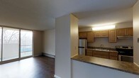 This single-floor unit offers a practical and functional layout with an open-concept design that connects the kitchen and living area seamlessly. The kitchen is defined by warm honey-toned wood cabinetry, dark countertops, a stainless steel refrigerator, and a double basin sink, all illuminated by overhead fluorescent lighting. A half-wall partition separates the kitchen from the adjacent living space, providing a sense of division while maintaining an open feel. The living area features dark laminate hardwood flooring that adds warmth and contrast to the neutral white walls. A large sliding glass door on the far wall allows natural light to filter into the space, brightening the interior and providing a view to the outdoors. A baseboard heating unit runs along the exterior wall beneath the window, ensuring climate comfort throughout the unit. The textured ceiling adds a subtle architectural detail consistent throughout the space. Overall, the unit presents a clean, straightforward layout well-suited for everyday living, with a balance of functional kitchen space and a comfortable open living area.