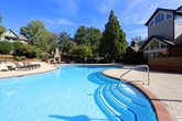 This single-story property features a striking combination of brick and wood exterior elements, complemented by mature, well-maintained landscaping at its entrance. A prominent chimney and large multi-pane windows add architectural character to the façade, while colorful potted flowers and sculpted shrubs create an inviting approach.

The centerpiece of the outdoor space is a generously sized freeform swimming pool with a distinctive brick-trimmed edge and vivid blue water. The expansive concrete pool deck provides ample space for lounging, furnished with multiple chaise lounges fitted with cushioned seating. A standout feature is the stone-clad outdoor fireplace, which serves as a focal point for a dedicated seating area ideal for gathering.

An integrated stone and brick retaining wall curves alongside the pool area, incorporating what appears to be an outdoor bar or kitchen space, topped with planters of vibrant flowers. The surrounding landscape is richly layered with mature deciduous trees that provide natural shade and privacy. The overall ambiance is resort-like, combining functional outdoor amenities with thoughtfully designed hardscaping and natural greenery throughout.