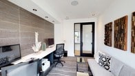 This single-floor space is thoughtfully designed to function as both a productive work environment and a comfortable lounge area. The layout is efficient yet stylish, balancing professional utility with contemporary design sensibility.

The workstation area is anchored by a built-in, floating desk finished in dark wood tones, set against a striking full-height tile accent wall in a warm gray-brown hue. The desk accommodates dual monitors and includes integrated storage below, providing ample workspace. Recessed lighting is positioned throughout the ceiling, ensuring consistent, even illumination across the room.

Opposite the workstation, a gray upholstered sofa with patterned accent pillows offers a relaxed seating option, paired with a sleek low-profile coffee table. A striped area rug grounds the central space, adding texture and visual interest underfoot.

A large picture window along one wall floods the interior with natural light while framing views of the surrounding outdoor landscape and adjacent buildings. Decorative wall art featuring carved wood and mosaic-style panels adds an artistic focal point. Black-framed glass doors provide a transparent connection to the adjoining corridor, maintaining an open and airy feel throughout the space.