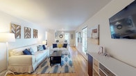 This single-floor home offers a well-organized layout with a cohesive, light-filled aesthetic throughout. Crown molding and warm hardwood flooring are consistent features that lend a classic, polished character to the space.

The living room is generously proportioned, with multiple windows along the exterior walls that invite abundant natural light and views of the surrounding greenery. The room comfortably accommodates two sofas arranged around a dark wood coffee table, complemented by a patterned area rug in blue and yellow tones. A storage unit and decorative wall art add functional and visual interest to the space.

The dining area, adjacent to the living room, features a round wooden table with seating for four, a pendant light overhead, and a window that provides additional natural light. It connects directly to the kitchen, which is equipped with white cabinetry, dark countertops, and white appliances including a range, over-the-range microwave, and refrigerator, alongside dark tile flooring.

The primary bedroom features multiple windows on two walls, creating a bright and airy atmosphere, with hardwood floors and ample closet access. The secondary bedroom is cozy and well-lit, with hardwood flooring and a window with garden views. The bathroom includes a full soaking tub, dark tile flooring, and a vanity with dark countertops.