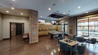 This single-floor amenity lounge is designed with a contemporary industrial aesthetic, blending exposed brick walls, polished concrete floors, and dark metal accents to create a dynamic and inviting social space. The main game area serves as the centerpiece, equipped with a billiards table illuminated by bold black pendant lights, wall-mounted flat-screen TVs, and a built-in wooden banquette seating wall. A full-length shuffleboard table is positioned along a dramatic black steel grid partition, which separates the space while allowing light and sightlines to pass through. Large windows and glass doors along one wall provide views of an outdoor pool area, flooding the interior with natural light. Comfortable seating clusters featuring teal upholstered chairs and cafe-style tables are distributed throughout, offering casual spaces for gathering. A dedicated kitchenette area features exposed brick backsplash, open dark shelving, a stainless steel sink, and a stainless dishwasher beneath a wood-beamed ceiling, complemented by a generous white countertop workspace. Recessed lighting throughout ensures the entire space is well-lit, while the cohesive use of brick, metal, and warm wood tones creates an engaging, club-like atmosphere suited for both socializing and recreation.