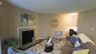 This single-story space in Irving, Texas, offers a welcoming living area centered around a sleek, tile-surround fireplace that serves as a natural focal point. The room is finished in warm, neutral tones — soft taupe walls paired with crisp white baseboards — creating a calm and inviting atmosphere. Dark hardwood-style flooring grounds the space and contrasts elegantly with the light-colored walls. A bold, geometric-patterned area rug defines the seating zone, adding visual interest and texture underfoot. The living area is thoughtfully arranged with a cushioned sofa, an accent chair in muted blue, and a contemporary glass-top coffee table. Decorative touches, including a sunburst mirror and an abstract canvas artwork in cool greens and blues, lend the space a curated, modern aesthetic. The curved wall transition between spaces adds an architectural detail that softens the overall layout. Ambient lighting warms the room, contributing to its comfortable, lived-in feel. The overall design balances practicality and style, making it a functional yet visually appealing single-floor living environment.