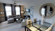 This single-floor space showcases an open-concept layout that seamlessly connects the living and dining areas, creating a cohesive and functional flow throughout. The living area is anchored by a plush grey sectional sofa paired with a sleek, rounded metallic coffee table, lending a modern and sophisticated feel. A ceiling fan overhead provides both practicality and a finishing architectural touch. Natural light filters through multiple windows dressed with blinds and dark curtain panels, illuminating the neutral grey walls and soft carpeted flooring. A console table along the wall serves as a display surface, adorned with decorative accents and framed artwork that reinforce the space's contemporary aesthetic. Abstract wall art in cool, silvery tones adds visual interest and depth to the room. The dining area features a glass-top table surrounded by cream and dark-framed chairs, complemented by a decorative round mirror on the wall. Throughout, the space maintains a cohesive color palette of greys, creams, and metallic accents, resulting in an elegant and well-appointed interior that balances comfort with modern style.