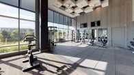 This single-level fitness center is designed to support a wide range of workout disciplines within a thoughtfully organized layout. The main exercise floor is defined by its impressive double-height ceilings, accented by a decorative coffered ceiling treatment, and bordered by expansive floor-to-ceiling windows that flood the space with natural light and offer open outdoor views. The flooring transitions between durable rubber tile in the main workout area and hardwood in the dedicated studio room, clearly delineating functional zones.

The primary gym floor is well-equipped with an extensive free weights section, including multiple racks of dumbbells and kettlebells of varying sizes, adjustable benches, and a selection of cable and resistance machines. Cardio equipment, including treadmills, ellipticals, and stationary bikes, is positioned to take advantage of the natural light from the large glazed window walls. Full-length mirrors along one wall visually expand the space and aid in form monitoring.

A separate studio room features light hardwood flooring, a wall-mounted flat-screen display, a built-in interior viewing window, and storage for stability balls and step platforms—ideal for group classes or individual training sessions. The overall aesthetic is clean, contemporary, and professional throughout.