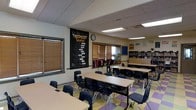 This elementary school classroom is thoughtfully arranged to foster an engaging and inclusive learning environment. The room is anchored by a bold chalkboard wall featuring the words "Learning Center," adorned with an uplifting message reminding students of their value and potential — setting a positive tone the moment anyone enters. Student desks and chairs are neatly organized in clusters, encouraging collaborative learning while allowing adequate space for movement throughout the room. A colorful, multi-patterned tile floor in shades of yellow, green, and purple adds a cheerful, vibrant energy to the space. Natural light filters in through several windows fitted with wooden blinds, complementing the overhead fluorescent panel lighting recessed into a drop ceiling. Along the back wall, well-stocked bookshelves offer accessible reading materials, while educational posters and bulletin boards display learning resources around the room. Backpacks and personal belongings are neatly stored along one wall, reflecting an organized and student-friendly setup. The overall ambiance is bright, orderly, and purpose-built to support active learning and student well-being.