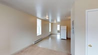 This single-floor space offers a straightforward and functional layout with a clean, neutral aesthetic throughout. The main living area is open and airy, featuring large double-hung windows that allow generous amounts of natural light to fill the room. The walls are finished in a soft, warm beige tone that complements the light-colored ceiling, giving the space a bright and welcoming feel.

Flooring transitions from carpet in the living area to a speckled hard floor surface near the kitchen zone, providing a practical distinction between the two areas. Baseboard heating units run along the exterior walls, ensuring efficient climate control throughout the space.

The kitchen area is positioned toward the rear of the unit and includes a full-size white refrigerator. A doorway leads to an additional room beyond, suggesting further utility or living space. An electrical panel is mounted on the wall near the kitchen, offering convenient access.

Flush-mount ceiling light fixtures illuminate the interior, contributing to a simple and uncluttered overhead profile. The overall ambiance of the space is modest and functional, with ample room to personalize and furnish according to individual needs. The open flow between the living and kitchen areas makes the space feel cohesive and easy to navigate.