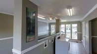 This single-floor laundry facility is designed with functionality and comfort in mind. The space features a row of full-sized washing machines and dryers positioned along a windowed wall, allowing abundant natural light to fill the room and offering views of the surrounding exterior. Large windows contribute to an open, airy feel throughout the area.

The interior is finished with neutral gray walls accented by crisp white crown molding and chair rail trim, lending a polished appearance to an otherwise utilitarian space. A textured ceiling adds dimension, while a ceiling fan with integrated lighting ensures adequate air circulation and illumination. Recessed display niches are built into the lower portion of the walls, adding a subtle architectural detail. A counter-height dividing partition provides a practical surface for sorting or folding laundry.

Overhead fluorescent lighting supplements natural light, ensuring the space remains bright at all times. Decorative wall art featuring a tropical waterfall scene adds a welcoming, homey touch to the environment. The tile flooring is easy to maintain and well-suited for the demands of a high-traffic laundry area. Overall, this facility balances practicality with thoughtful design details to create a comfortable and efficient shared laundry experience.