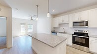 This single-floor, one-bedroom, one-bathroom unit offers a thoughtfully designed layout that maximizes functionality within a compact footprint. The open-concept kitchen and living area forms the heart of the unit, anchored by a substantial granite-topped island that provides generous prep and dining space. The kitchen is outfitted with white shaker-style cabinetry, a tile backsplash, and a full suite of stainless steel appliances, including a refrigerator with ice and water dispenser, electric range, over-the-range microwave, and dishwasher. Light wood-look flooring runs throughout the main living area, complementing the bright, neutral palette. Pendant lighting over the island and a ceiling fan in the living room add functional style, while a glazed exterior door draws in natural light and provides outdoor access. The bedroom features plush carpeting, a ceiling fan, and a window overlooking greenery, creating a calm retreat. The bathroom includes a granite-topped vanity, wood-look flooring, and accessibility grab bars. A dedicated laundry room houses a full-size Whirlpool washer and dryer with overhead wire shelving for added storage.