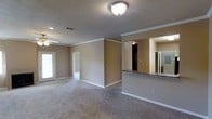 This single-floor residence showcases an open-concept layout that seamlessly connects the living area to the kitchen. The spacious main living area is finished with plush wall-to-wall carpeting and neutral taupe-toned walls, creating a warm and inviting atmosphere. Crown molding runs along the ceiling perimeter, adding a refined architectural detail throughout the space. A ceiling fan is visible in the living area, contributing to both comfort and airflow.

Natural light enters through a window along the far wall, brightening the open floor plan. A doorway leads to an additional room, likely a bedroom or hallway, extending the functional layout of the home.

The kitchen is defined by a raised breakfast bar counter finished with dark granite-style countertops, which serves as a natural dividing element between the living and kitchen spaces. The pass-through design maintains an open sightline between rooms. A mirrored surface and refrigerator are visible within the kitchen area, hinting at a well-equipped cooking space. Textured ceilings, white baseboards, and consistent neutral tones throughout unify the overall design aesthetic.