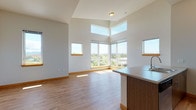 This well-designed single-floor residence offers a thoughtful layout with a clean, contemporary aesthetic throughout. The open-concept living and dining area is a standout feature, boasting soaring ceilings with elevated clerestory windows that flood the space with natural light. A dramatic corner window arrangement frames sweeping panoramic views of the surrounding landscape and treetops, creating a bright and airy atmosphere. Warm wood-tone baseboards and durable hardwood-style flooring flow throughout the main living areas, adding a welcoming feel.

The kitchen is efficiently designed with a large center island featuring an integrated sink and generous countertop space finished in a neutral tone. Cabinetry in a warm wood grain finish lines the walls, complemented by stainless steel appliances including a refrigerator, range, and over-the-range microwave.

The bedrooms are carpeted for comfort, each offering access to natural light through well-placed windows with scenic outdoor views. The primary bedroom features soaring ceilings, a sliding-door closet, and a large window. The full bathroom includes hardwood-style flooring, a combined tub and shower, a vanity with storage, and modern fixtures. Light wood doors and trim carry a consistent, cohesive design language throughout the entire space.
