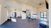 This single-floor fitness center is thoughtfully designed to support a wide range of workout styles and training needs. The space is divided into distinct functional zones, seamlessly connected through open archways and structural columns that define the layout without closing off the floor plan.

The strength and free weights area is well-appointed with a full dumbbell rack, medicine balls, stability balls, a weight bench, a power rack with barbell, and a hanging heavy bag — all positioned on durable dark rubber flooring. Motivational wall graphics, including bold "Harder. Better. Faster. Stronger." and "Hustle & Muscle" lettering, energize the environment. A large wall-mounted mirror amplifies the sense of space and aids in form training.

The cardio zone is flooded with natural light through expansive floor-to-ceiling sliding glass doors that open to a view of a resort-style pool and outdoor amenity area. Cardio equipment includes a treadmill, rowing machine, elliptical, and stationary bike, all arranged to take advantage of the outdoor views. A wall-mounted flat-screen TV and ceiling fans add comfort and convenience throughout.

Crown molding and recessed lighting throughout lend a refined, polished character to the space, elevating it well beyond a standard fitness facility.