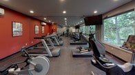 This fitness center is thoughtfully designed to support a comprehensive workout experience within a single, open floor layout. The space features a bold design aesthetic, with deep red accent walls contrasting against neutral gray walls and ceilings, creating an energizing atmosphere. Recessed lighting throughout ensures the room is well-lit, complemented by large windows along one wall that frame serene views of mature trees and natural greenery, bringing the outdoors in and enhancing the workout environment.

The cardio zone is generously outfitted with a row of Matrix treadmills, elliptical trainers, and stationary bikes, including a recumbent cycle. Wall-mounted flat-screen televisions are positioned for easy viewing from the cardio equipment. A full-length mirror spans one wall, amplifying the sense of space and allowing users to monitor their form. The strength and functional training area includes a multi-use cable machine, weight bench, stability balls, and kettlebells, providing versatility for a range of training routines.

The flooring consists of durable rubber sport surfacing, practical for both cardio and strength activities. A row of private changing rooms or storage enclosures with glass-paneled doors lines one wall, adding a functional and organized element to the overall layout. The space is efficiently arranged to maximize usability while maintaining an open, uncluttered feel throughout.