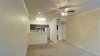 This single-floor residence showcases an open-concept design that seamlessly connects the living area and kitchen, creating a spacious and welcoming atmosphere. The living room is finished with wall-to-wall carpeting and features a multi-blade ceiling fan with an integrated light fixture, providing both comfort and ambient lighting. Crown molding trims the ceiling throughout, adding a refined architectural touch to the space.

The kitchen is positioned adjacent to the living area and is defined by a raised breakfast bar that serves as a natural divider between the two spaces. Dark espresso cabinetry contrasts elegantly with the light-toned walls, while stainless steel appliances — including a refrigerator, microwave, and range — lend a modern, polished look. A tile backsplash adds texture and detail to the cooking area.

A hallway visible from the main living space leads to additional rooms, offering a sense of depth and privacy beyond the open living zone. Recessed and overhead lighting ensures the kitchen remains bright and functional. The neutral color palette of warm whites and beiges throughout creates a cohesive, inviting ambiance well-suited for everyday living.