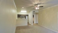 This single-floor residence showcases an open-concept design that seamlessly connects the living area and kitchen, creating a spacious and welcoming atmosphere. The living room is finished with wall-to-wall carpeting and features a multi-blade ceiling fan with an integrated light fixture, providing both comfort and ambient lighting. Crown molding trims the ceiling throughout, adding a refined architectural touch to the space.

The kitchen is positioned adjacent to the living area and is defined by a raised breakfast bar that serves as a natural divider between the two spaces. Dark espresso cabinetry contrasts elegantly with the light-toned walls, while stainless steel appliances — including a refrigerator, microwave, and range — lend a modern, polished look. A tile backsplash adds texture and detail to the cooking area.

A hallway visible from the main living space leads to additional rooms, offering a sense of depth and privacy beyond the open living zone. Recessed and overhead lighting ensures the kitchen remains bright and functional. The neutral color palette of warm whites and beiges throughout creates a cohesive, inviting ambiance well-suited for everyday living.