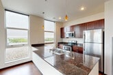 This thoughtfully designed one-bedroom, one-bathroom residence offers a comfortable and modern living experience across a single floor. The open-concept kitchen and living area forms the heart of the space, where rich hardwood-style flooring flows throughout and abundant natural light pours in through expansive floor-to-ceiling windows. The kitchen is a standout feature, showcasing deep-toned cabinetry, dark granite countertops, a large center island with a double sink, and a full suite of stainless steel appliances including a refrigerator, range, and built-in microwave. Decorative pendant lighting adds a warm, stylish accent above the island. The living area is generously proportioned, with large sliding glass doors opening to an outdoor balcony and offering panoramic views of the surrounding cityscape and natural landscape beyond. The bedroom is a serene retreat, carpeted for comfort and bathed in natural light from oversized windows that continue the space's connection to the outdoors. A dedicated closet provides practical storage. The bathroom is cleanly finished with a rich wood vanity, stone-toned countertop, tile flooring, and a full tub-shower combination. Throughout the residence, the consistent use of quality materials and modern finishes creates a cohesive and inviting atmosphere.