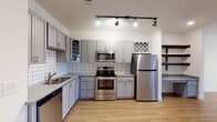 Located on W Kinderton Way in Bermuda Run, NC, this single-floor unit offers a thoughtfully designed layout with clean, modern finishes throughout. The kitchen is a standout feature, showcasing gray shaker-style cabinetry, granite countertops, a white subway tile backsplash, and a full suite of stainless steel appliances including a refrigerator, electric range, built-in microwave, and dishwasher. Track lighting illuminates the space, and open wood shelving adds a warm, functional accent. The kitchen flows seamlessly into a generous open living area finished with light hardwood flooring that extends throughout the main space, creating a bright and cohesive feel. French doors at the far end of the living area invite natural light and provide access to an outdoor balcony. The bedroom is carpeted for comfort and includes a ceiling fan and a window that brings in additional natural light. A spacious walk-in closet with wire shelving systems offers ample storage. The bathroom features a tub-shower combination with white tile surround, granite-topped vanity with gray cabinetry, an overhead storage cabinet, and built-in wood shelving for added organization. Light hardwood flooring ties the bathroom together with the rest of the unit's design aesthetic.