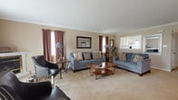 This single-floor home offers a thoughtfully arranged layout that balances comfort and functionality across its key living spaces. The living room serves as the centerpiece, featuring warm neutral walls, crown molding, and plush carpeting. Multiple windows dressed in rich burgundy curtains allow natural light to fill the room, while a fireplace adds a classic focal point. Ample seating space makes it well-suited for both relaxing and entertaining.

The kitchen is outfitted with white cabinetry, dark countertops, and light hardwood flooring, providing a clean and practical workspace. White appliances including a range and built-in microwave are neatly integrated. An adjacent dining nook with large windows and hardwood flooring offers a bright, casual setting for meals.

The home includes two bedrooms, each carpeted and finished with neutral tones. The primary bedroom features a ceiling fan, natural light from a window, and generous closet access. The second bedroom is distinguished by a calming blue accent wall. Both bathrooms are finished in a clean white palette with vanity cabinetry, wall-mounted mirrors, built-in shelving, and tile flooring, providing efficient and functional spaces throughout.