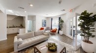 This versatile space showcases a seamless blend of comfort and functionality. The open-concept living area boasts hardwood floors, recessed lighting, and a neutral color palette, accented by pops of vibrant orange in the furniture. A state-of-the-art fitness center features cardio equipment, free weights, and a dedicated yoga space with modern geometric carpeting. The kitchen area impresses with sleek white cabinetry, stainless steel appliances, and a unique mountain-inspired backsplash. Large windows throughout provide ample natural light, while strategically placed indoor plants add a touch of nature to the contemporary setting. The space is designed for both relaxation and productivity, with multiple seating areas and a cohesive, modern aesthetic.