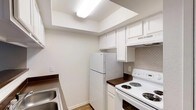 This single-floor space includes a kitchen designed for everyday functionality. The kitchen features white upper and lower cabinetry that provides ample storage, complemented by dark countertops that offer a subtle contrast. A full-size white refrigerator and a white electric coil-burner range anchor the layout, with a built-in range hood overhead. A stainless steel sink is positioned along the adjacent wall, adding a practical touch to the workspace. The backsplash behind the range showcases a neutral-toned brick-pattern tile, introducing a layer of texture to the otherwise clean, minimalist aesthetic. Overhead, a recessed ceiling detail with fluorescent lighting brightens the space effectively. The light-toned flooring runs throughout, contributing to the open and airy feel of the kitchen. The overall color palette — white cabinetry, neutral walls, and soft flooring — creates a cohesive and well-lit environment. The layout is compact yet efficiently arranged, making good use of the available space for both storage and meal preparation.