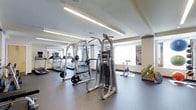Located at 1 Cotton St in the Manayunk neighborhood of Philadelphia, PA, this fitness center offers a well-appointed, single-floor workout facility designed to support a comprehensive exercise routine. The space is outfitted with a variety of strength training equipment, including cable machines and weight racks, alongside cardio machines such as treadmills and stationary bikes. Large mirrors line the walls, enhancing the sense of openness and allowing users to monitor their form during workouts. Natural light filters in through windows along the perimeter, complemented by sleek recessed linear lighting fixtures mounted on the clean white ceiling, creating a bright and energizing atmosphere. The flooring features durable, dark rubberized material well-suited for a high-use fitness environment. A decorative accent wall adds a touch of color and visual interest to the otherwise modern, minimalist aesthetic. The layout is thoughtfully organized to provide ample space between equipment, ensuring a comfortable and safe workout experience. Overall, the facility presents a clean, modern environment equipped to meet the needs of a wide range of fitness enthusiasts.