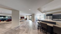 This thoughtfully designed single-floor amenity space offers a seamless blend of entertainment and relaxation zones, unified by a striking herringbone-patterned wood-look tile floor that flows throughout the open layout. The space is connected yet organized into distinct functional areas, each with its own character and purpose.

The wet bar and kitchenette area serves as a central hub, outfitted with navy blue cabinetry, quartz countertops, a stainless steel refrigerator, built-in microwave, and a subway tile backsplash with under-cabinet LED accent lighting. Open shelving adds both style and practicality to the space.

Adjacent to the kitchen, a spacious open area accommodates a pool table and shuffleboard, bathed in natural light from large windows that offer outdoor views. The game room also features colorful artwork and a built-in niche with seating, contributing a lively, social atmosphere.

A dedicated media lounge provides a more intimate retreat, featuring plush red and burgundy seating, a large wall-mounted television, navy built-in cabinetry with LED cove lighting, and bold abstract artwork. French doors offer direct outdoor access, enhancing the overall sense of openness throughout the space.