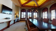 This sophisticated conference room blends traditional architectural design with functional meeting space. The room's most striking feature is its coffered ceiling, highlighted by vibrant yellow inlays and rich exposed wood beams that lend warmth and grandeur to the space. A wrought-iron chandelier with candle-style lighting hangs prominently above the elongated dark wood conference table, which is surrounded by comfortable upholstered rolling chairs in a warm terracotta hue.

A row of arched windows fitted with rich mahogany-stained wood shutters lines one wall, allowing natural light to filter in while offering outdoor views and privacy control. The carpet flooring in a neutral gold tone complements the warm wood tones found throughout the room's trim, baseboards, and door casings.

A built-in credenza with accent lamps runs along one wall alongside a wall-mounted flat-screen television, providing both practical workspace and presentation capabilities. An arched solid wood door and a set of glass-paned French doors with mahogany framing offer elegant entry points into the space.

The overall ambiance is refined and professional, combining a classic Mediterranean-inspired aesthetic with the functionality needed for executive meetings and collaborative gatherings.