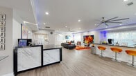 Located at 6115 N Davis Hwy in Pensacola, Florida, this single-floor commercial facility combines contemporary design with versatile, well-appointed spaces. The reception area makes a strong first impression with a striking marble-paneled front desk, light wood-look flooring, vibrant large-format artwork, and an accent-lit tray ceiling that casts a modern blue ambient glow. The open main area flows seamlessly from the reception into a stylish lounge and meeting space, featuring a glass-top conference table with sleek chrome-legged chairs, a wall-mounted display, large ceiling fans, and bold statement art. A bar-height counter with contemporary orange seating adds a dynamic social element. The fully equipped exercise room is a standout, offering rubber flooring, multiple cardio machines, strength equipment, ceiling fans, and ample natural light from large windows. An adjoining bonus room serves as a bright, cheerful children's play area with a wall-mounted TV, colorful rugs, and a direct interior window overlooking the fitness space. Throughout the facility, recessed lighting, crown molding, and a cohesive neutral palette create a polished, professional atmosphere that is both energetic and welcoming.