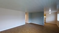 This single-floor residence presents a straightforward and functional layout designed for comfortable everyday living. The main living area is finished with warm hardwood-style flooring that extends throughout, providing a cohesive and inviting foundation for the space. Walls are painted in a combination of soft neutral tones — a crisp off-white on one side and a muted blue-gray accent wall on the other — creating a calm, understated ambiance. A textured ceiling adds subtle visual interest overhead. The living area is generously proportioned, offering ample room for a variety of furniture arrangements. Floor vents indicate a central HVAC system for year-round climate control, while multiple electrical outlets are conveniently positioned along the baseboards. An open passageway connects the living area to the adjacent kitchen, which features warm wood-toned cabinetry and appears to include countertop workspace and standard appliances, offering a practical cooking environment. The open sightlines between the living and kitchen areas contribute to an airy, connected feel throughout the home. White baseboards frame each room cleanly, tying together the overall aesthetic. The space is vacant and move-in ready, making it easy to envision a personalized interior arrangement.