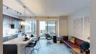 This single-floor residence showcases a thoughtfully designed open-concept layout that integrates the dining and living areas into one cohesive, light-filled space. Large floor-to-ceiling windows dominate the exterior wall, flooding the interior with abundant natural light while offering views of the surrounding building exterior and beyond. Sliding glass doors lead directly to a private balcony, blurring the boundary between indoor and outdoor living.

The dining area features a sleek, white countertop bar with modern black bar stools positioned beneath a minimalist pendant light fixture, creating a clean and contemporary aesthetic. Light hardwood-style flooring runs throughout, contributing to the airy, open feel of the space.

The living area is anchored by a rich, tufted leather sofa accented with vibrant patterned throw pillows, layered over a soft blue area rug. A stylish accent chair and small side table complete the seating arrangement, adding functional elegance. Neutral wall tones and white ceilings enhance the overall brightness. The space balances modern design with warm, inviting furnishings, creating a comfortable yet sophisticated atmosphere.