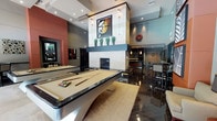 This single-floor entertainment lounge is designed with a contemporary aesthetic, blending bold design elements with functional recreational spaces. At the heart of the room sits a sleek billiards table with a cream felt surface, complemented by a polished dark floor that reflects the ambient lighting above. A dramatic stone fireplace serves as a central architectural focal point, framed by a tall column structure adorned with decorative artwork, wall sconces, and potted ornamental grasses on the mantle. The ceiling features recessed lighting and layered tray details, adding depth and dimension to the space. Accent walls in deep red and neutral gray tones create a dynamic visual contrast throughout. Toward the rear, a well-appointed bar area is visible, complete with dark wood cabinetry, pendant lighting, and high bar seating, making it ideal for socializing. A dartboard mounted on the wall adds a further recreational element. The overall ambiance is upscale yet inviting, combining polished surfaces, curated artwork, and thoughtful lighting to create a sophisticated entertainment environment suited for leisure and gatherings.