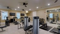 The fitness center at 2945 Midtown Way in Wilmington is a well-appointed, single-floor workout facility designed to support a range of exercise routines. The space features a clean, neutral interior with warm beige walls, crown molding, and light flooring that creates a bright and welcoming atmosphere. Recessed lighting throughout the ceiling ensures consistent illumination across the entire floor, complemented by ceiling fans that promote air circulation during workouts. Natural light filters in through multiple windows fitted with blinds, adding to the open feel of the space. The facility is equipped with Life Fitness cardio machines, including treadmills and an elliptical, alongside strength-training cable machines and a dedicated free-weight area featuring a fully stocked dumbbell rack. Flat-screen televisions are mounted on the walls near the cardio equipment, allowing residents to stay entertained during their workouts. A mirrored wall section enhances the sense of space while providing visual feedback for strength training. The layout is thoughtfully organized to allow comfortable movement between equipment stations, making the center practical and accessible for everyday use.