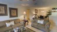 Located at 350 Byram Drive in Byram, Mississippi, this single-story residence offers a welcoming interior designed with a traditional aesthetic. The open-concept layout seamlessly connects the living room and dining area, creating a spacious and cohesive flow throughout the main living spaces. The living room is furnished with plush, neutral-toned sofas accented by decorative throw pillows, anchored by a patterned area rug and a glass-topped coffee table. A classic side table with an ornate base and a tall table lamp adds a refined touch, while framed wall art contributes warmth and character to the space. Visible wall vents indicate a central HVAC system throughout. The adjoining dining area is illuminated by an elegant chandelier, casting a warm glow over the dining table and dark upholstered chairs, making it well-suited for both everyday meals and entertaining. The light-colored flooring enhances the brightness of the interior, and the neutral wall tones provide a versatile backdrop. Overall, the home presents a comfortable, traditionally styled living environment with practical and cohesive design elements throughout.