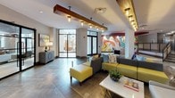 This contemporary lobby is designed to make a strong first impression, blending artistic expression with thoughtful architectural detail. Large-format stone-look tile flooring extends throughout, providing a cohesive and durable foundation for the open layout. A striking, large-scale abstract mural rendered in vivid reds, oranges, and yellows serves as a bold focal point, anchoring one end of the space with visual energy.

Suspended wood beam light fixtures run along the ceiling, adding warmth and an industrial-chic contrast to the otherwise crisp white walls and recessed lighting above. The lobby is divided into distinct zones: a main lounge area furnished with modular gray and yellow upholstered seating arranged around marble-topped tables, and a more intimate seating nook defined by a patterned area rug, vibrant accent chairs, and direct views to an outdoor courtyard through floor-to-ceiling black-framed windows.

A set of interior stairs with sleek black metal railings leads to a lower-level area, partially visible through glass-walled enclosures that house what appears to be a club room or amenity space. Structural white columns with textured paneling frame the transitions between zones, while natural light from the courtyard-facing glazing brightens the overall ambiance, creating a dynamic and inviting communal environment.