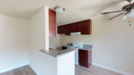 This single-floor space showcases a well-appointed kitchen as its central feature. The kitchen is defined by rich, dark mahogany-toned cabinetry that extends to the upper walls, providing generous storage capacity. The cabinetry is complemented by granite-style countertops in a neutral gray tone, offering a clean and cohesive aesthetic. A white electric range with overhead ventilation is seamlessly integrated into the cabinetry layout, contributing to the kitchen's practical design.

A peninsula counter with a white base and matching granite-style surface extends outward, creating a natural partition between the kitchen and the adjacent living area. This feature adds both additional prep space and a casual dining or gathering area. The light hardwood-style flooring runs throughout, lending warmth and continuity to the space.

Walls are finished in a soft neutral tone, enhancing the brightness of the room. A flush-mount ceiling light fixture provides functional overhead illumination. The textured ceiling adds a subtle architectural detail. Overall, the space offers a clean, move-in-ready environment with a practical and cohesive interior design.