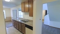 This two-story residence offers a practical and well-organized layout across its two levels. The kitchen serves as a central feature, showcasing warm-toned wood cabinetry both above and below the counters, providing ample storage throughout. The countertops display a speckled granite-style surface that adds a touch of durability and visual interest to the space. A double stainless steel sink is positioned along the counter run, accompanied by a built-in dishwasher, making the kitchen well-equipped for everyday use. A window positioned above the sink area allows natural light to filter into the space, offering a view to the exterior. The kitchen connects openly to the adjacent living areas, creating a cohesive flow throughout the main level. Light-toned tile flooring runs through the kitchen, transitioning to carpeted flooring visible in the neighboring rooms. A staircase is visible from the kitchen area, providing access to the second floor of the residence. The overall ambiance is neutral and functional, with a clean, understated aesthetic defined by warm wood tones and light wall colors throughout.